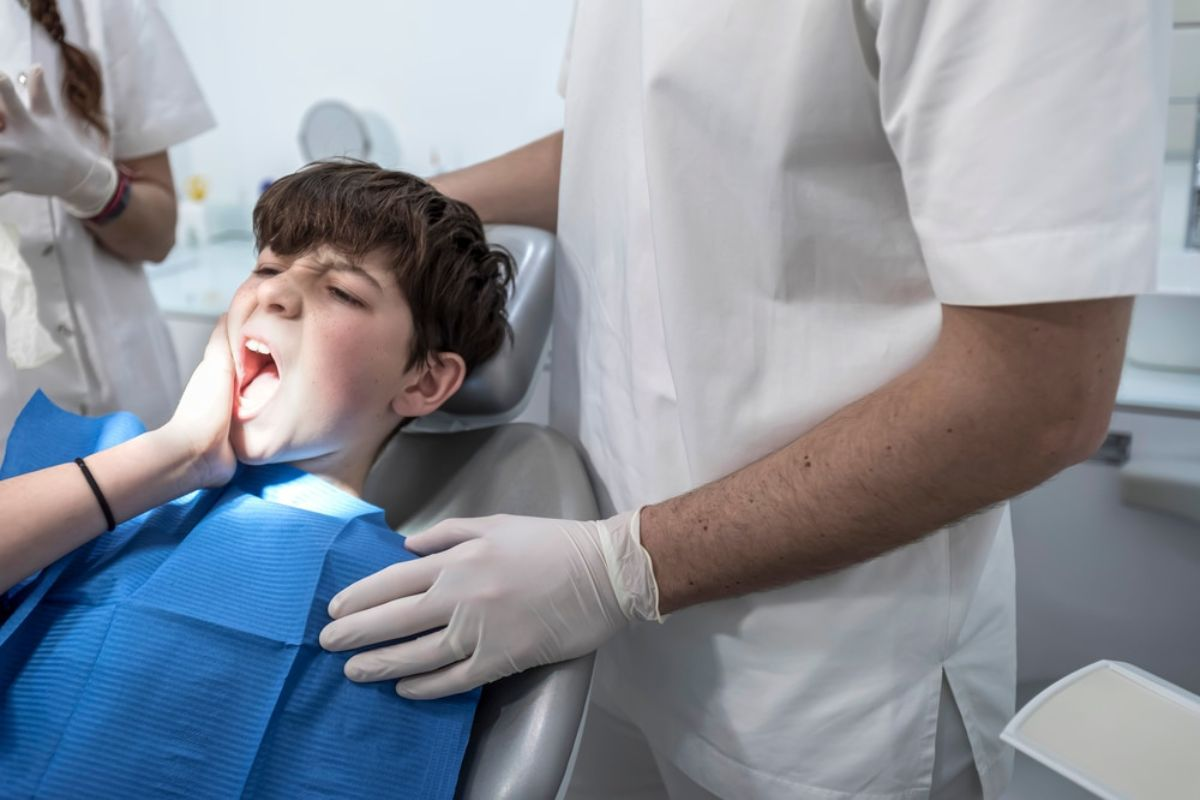 Boy with tooth pain sitting in dentist chair holding jaw while dentist with gloves comforts him.