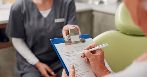Person filling out a medical form on a clipboard while sitting opposite a healthcare worker in scrubs.