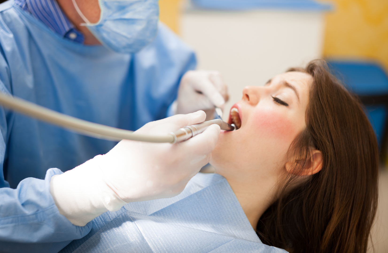 Dentist in blue scrubs and mask examining a woman's open mouth with dental tools.