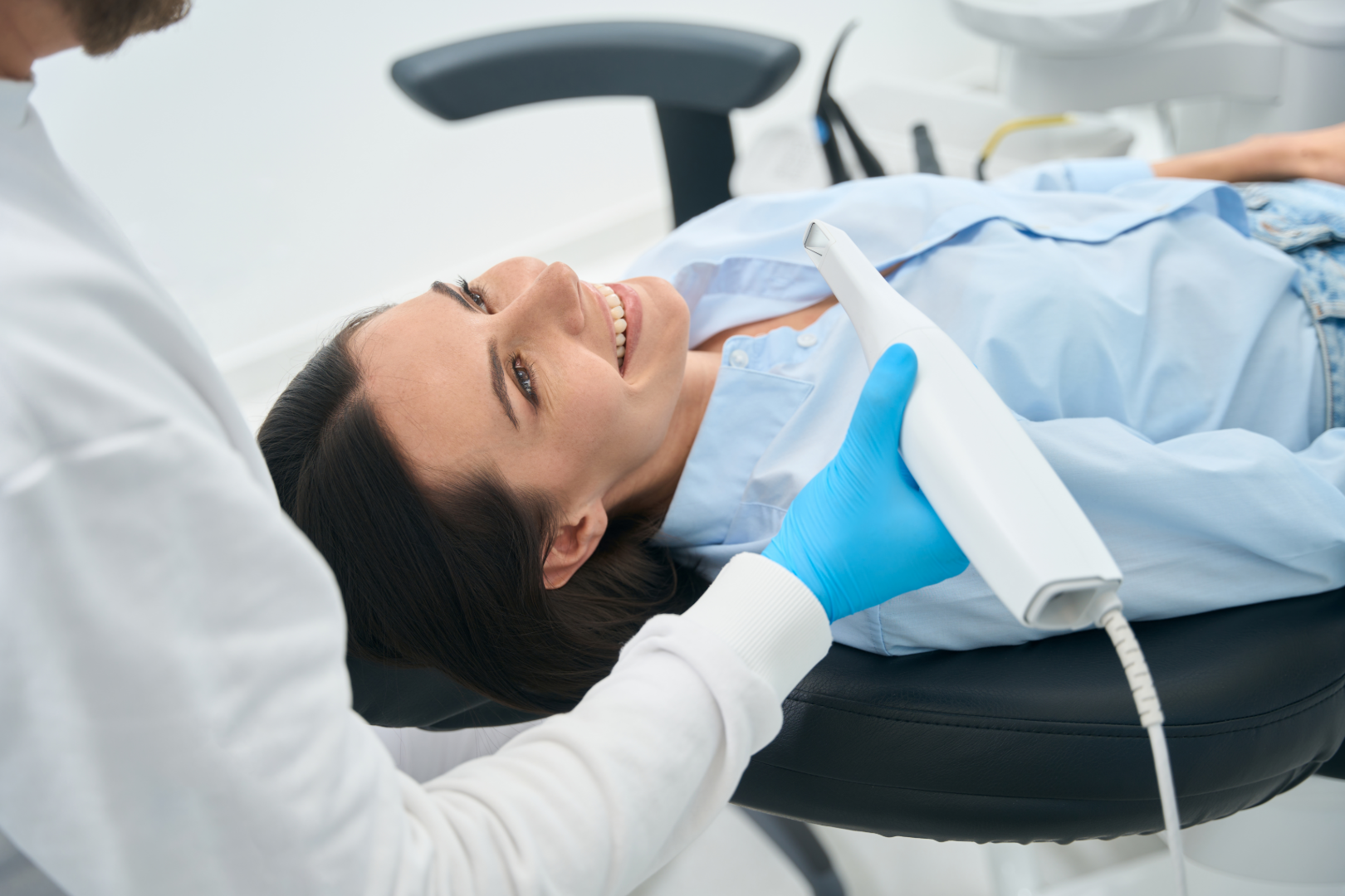 Smiling woman lying in dental chair while dentist wearing blue gloves holds an oral scanner.