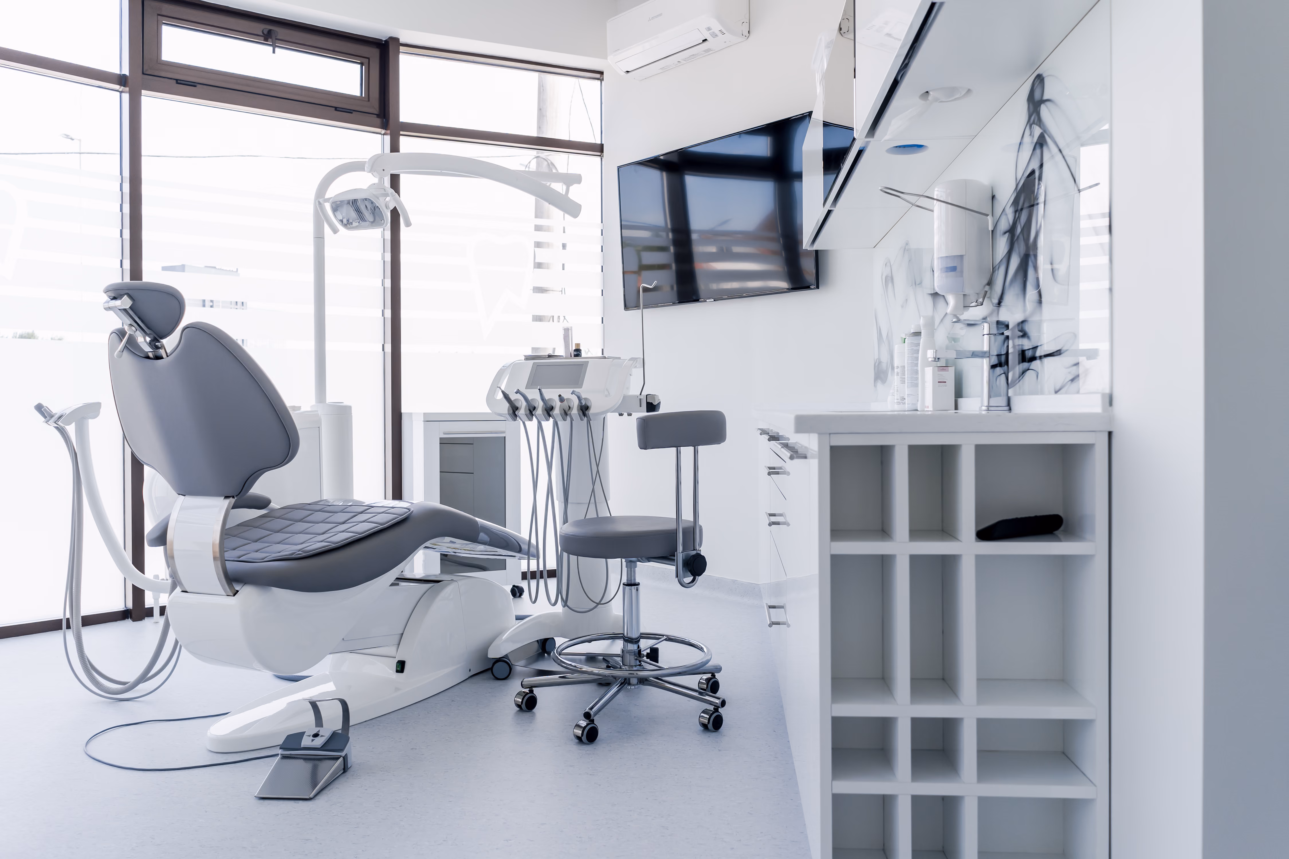 Modern dental office featuring patient chair, tools, stool, TV, and clean white cabinetry.