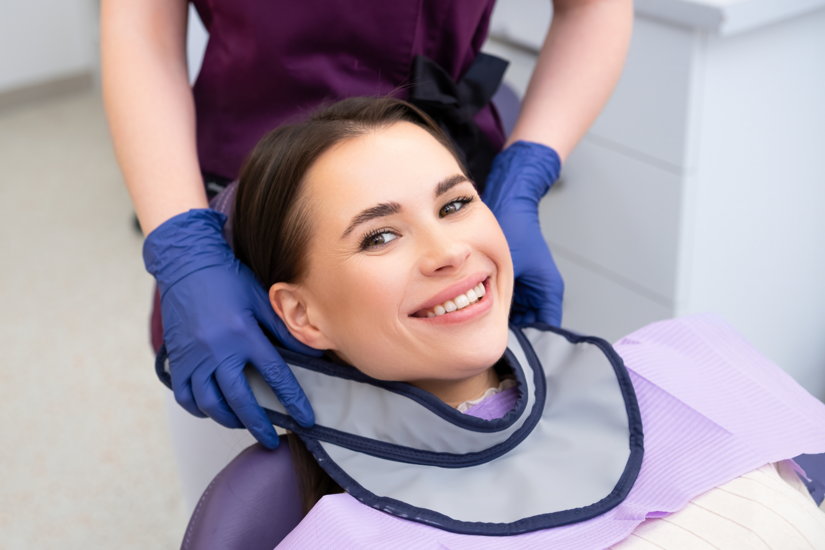Smiling woman in dental chair with a protective lead apron being placed by a gloved dental professional.