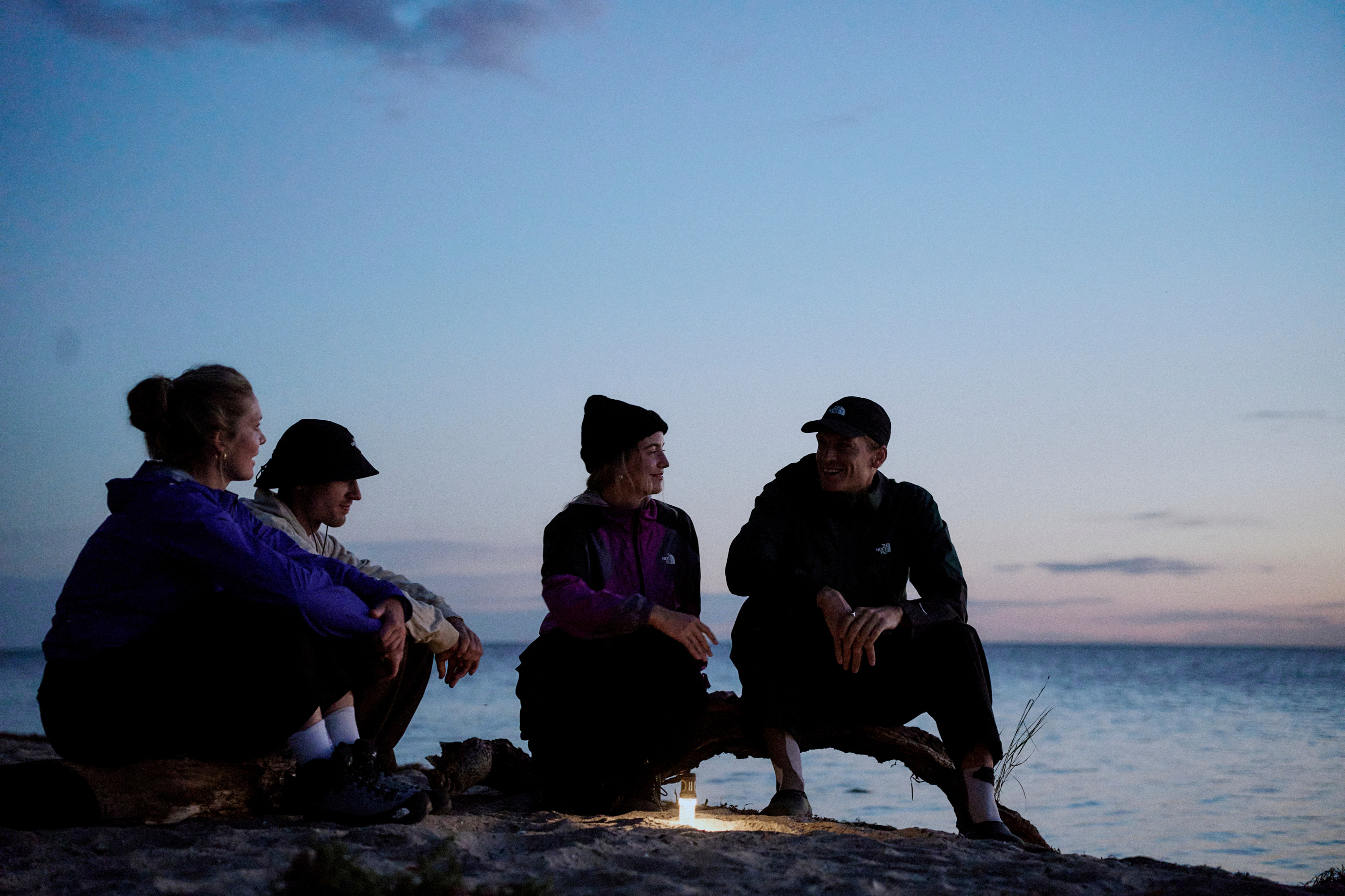 Four friends in The North Face jackets sitting by the coast at blue hour.