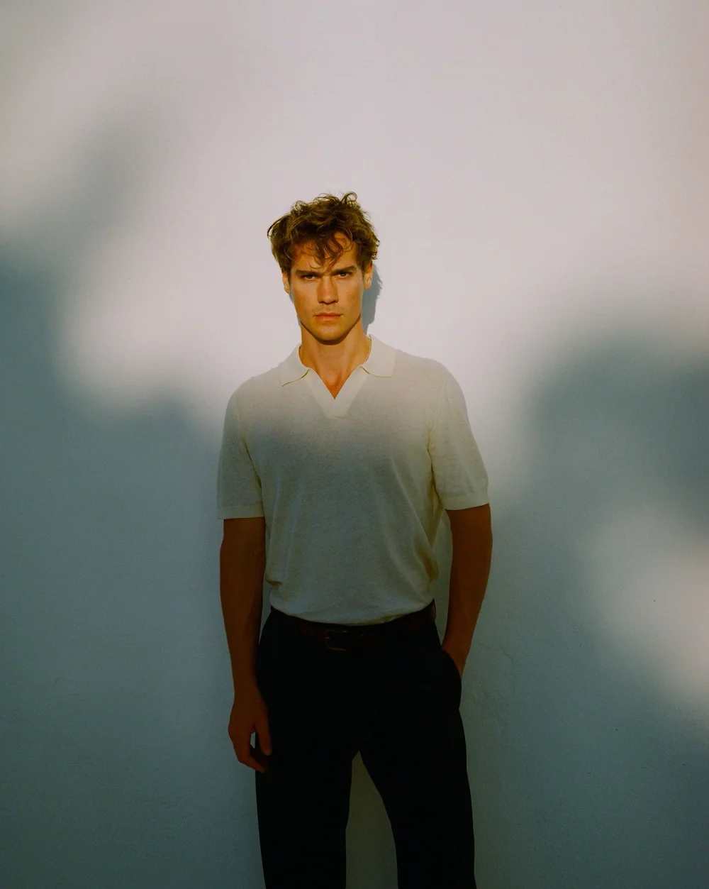 Patrick Pilz wearing a white polo shirt and dark trousers against a softly shadowed wall.