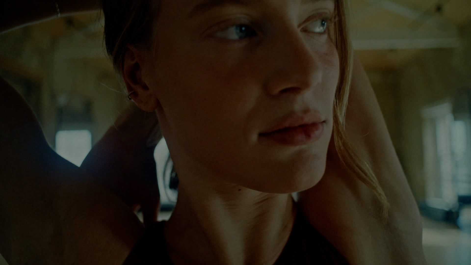 Close-up of a dancer looking to the side with her hands behind her neck in a sunlit studio.