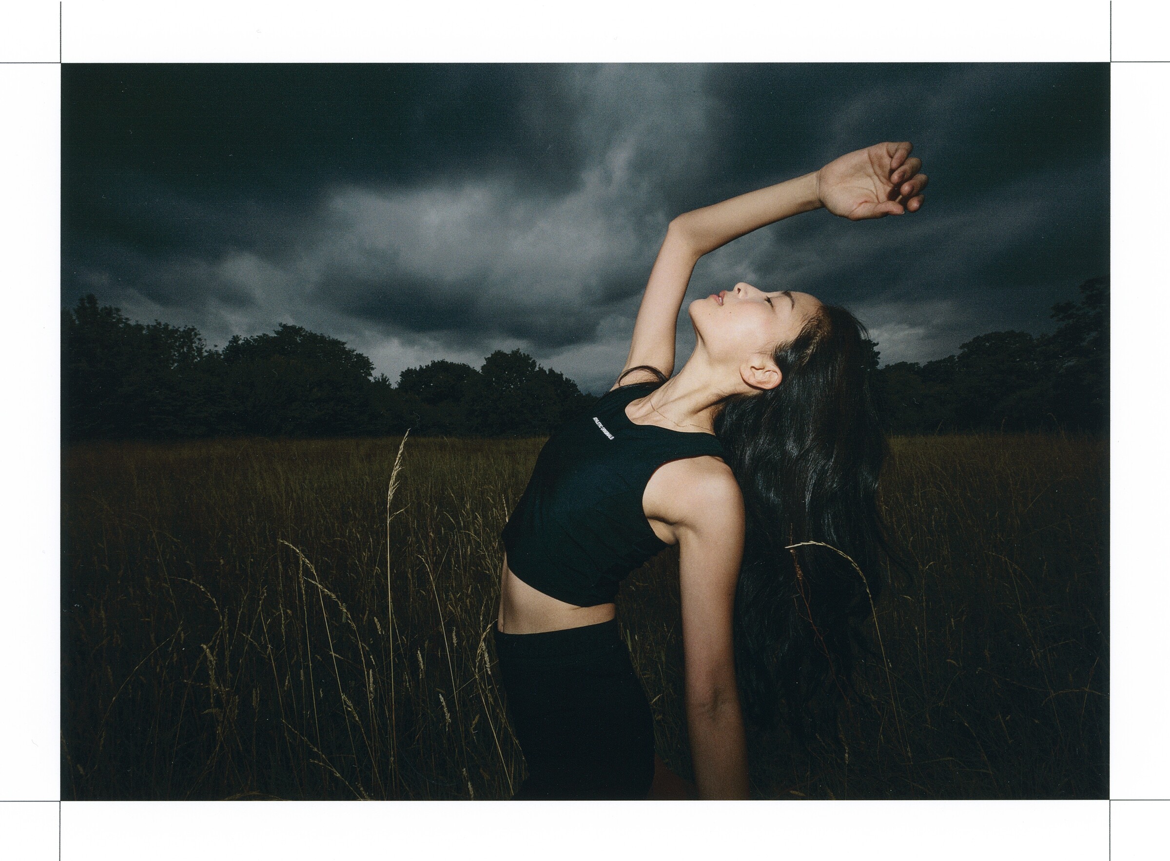 DESCENTE Athletic Originals: Woman stretching one arm overhead in tall grass under dark clouds.