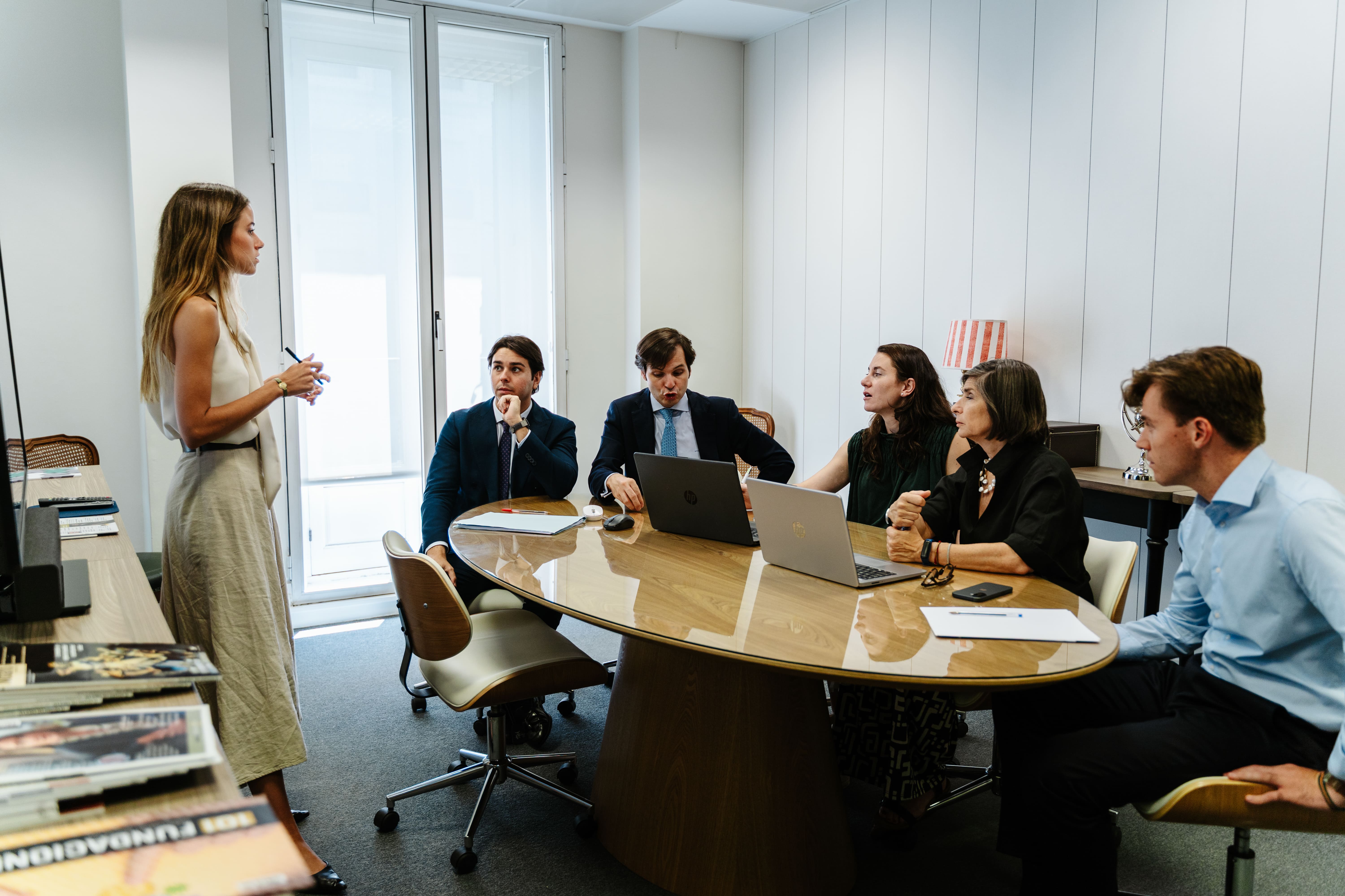 Una mujer de pie presenta a cuatro personas sentadas en una reunión de oficina junto a una mesa ovalada.