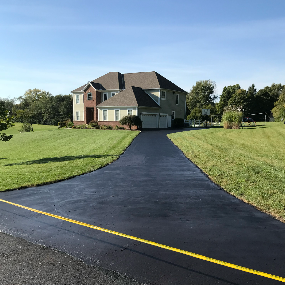 Long residential asphalt driveway after sealcoating, leading to a two-story brick home
