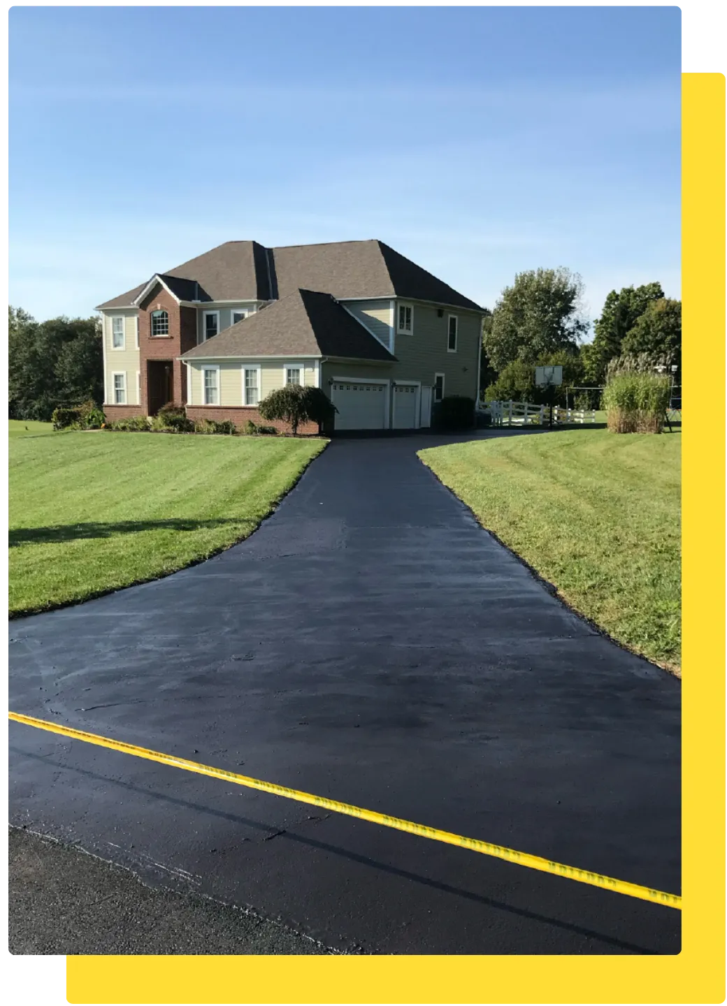 Long residential asphalt driveway after sealcoating, leading to a two-story brick home