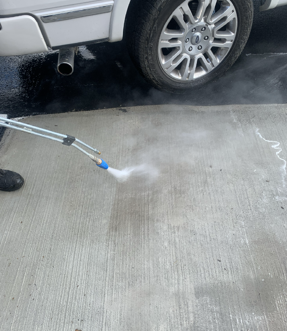 Pressure washing a driveway surface beside a parked car before sealcoating