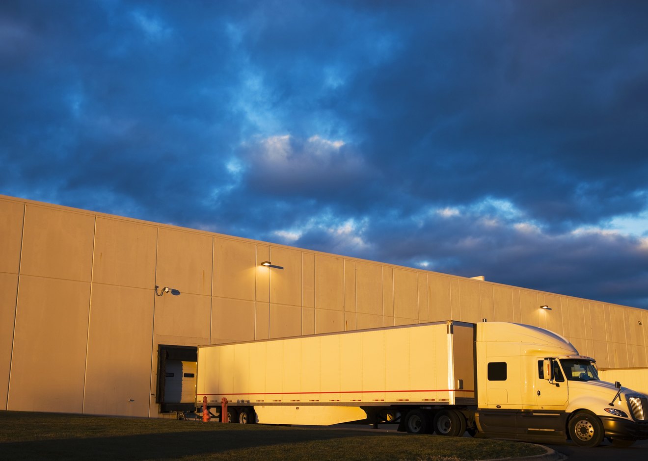 A tractor-trailer backed into a warehouse loading dock at dusk, low sun on the building wall.