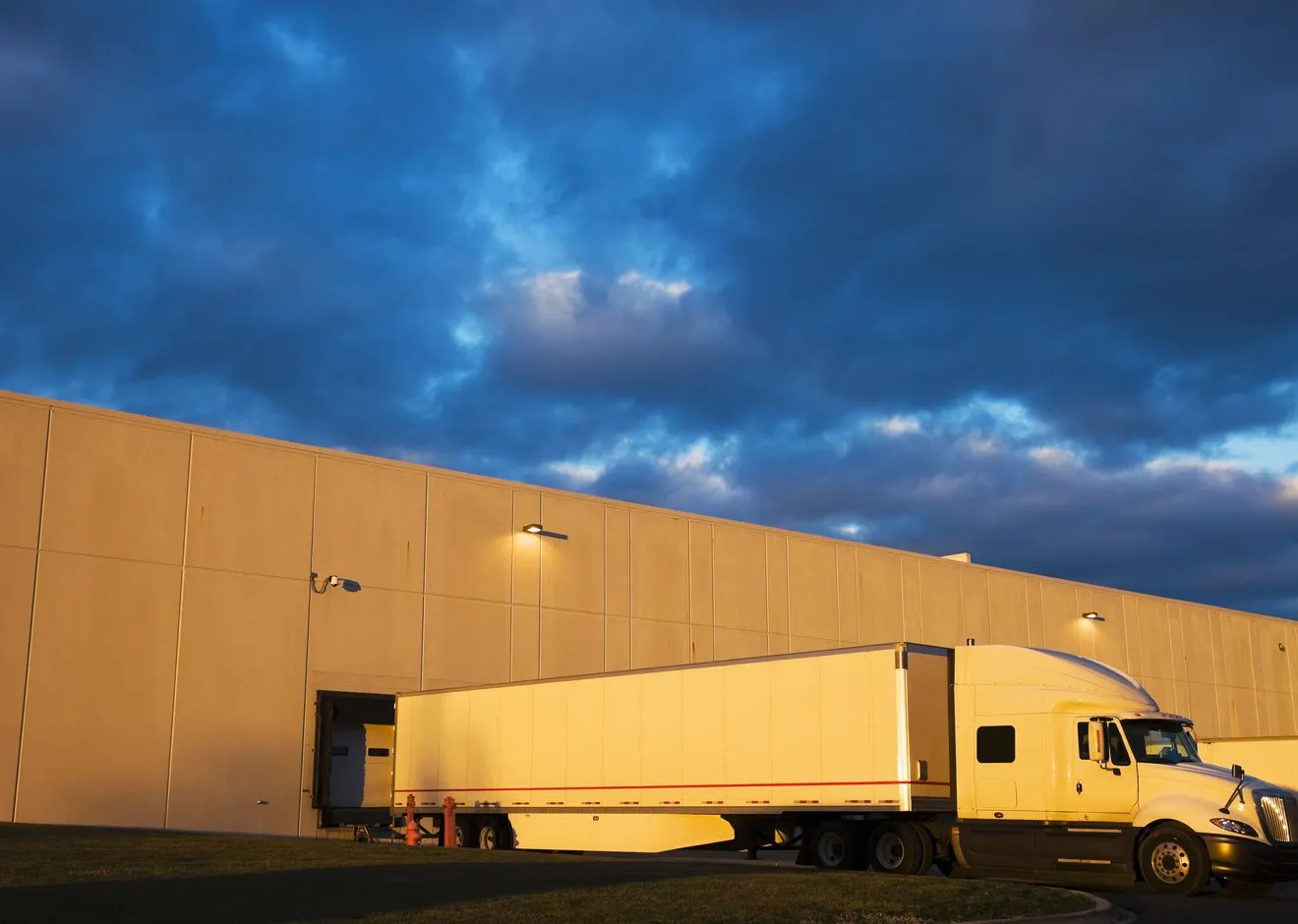 A tractor-trailer backed into a warehouse loading dock at dusk, low sun on the building wall.