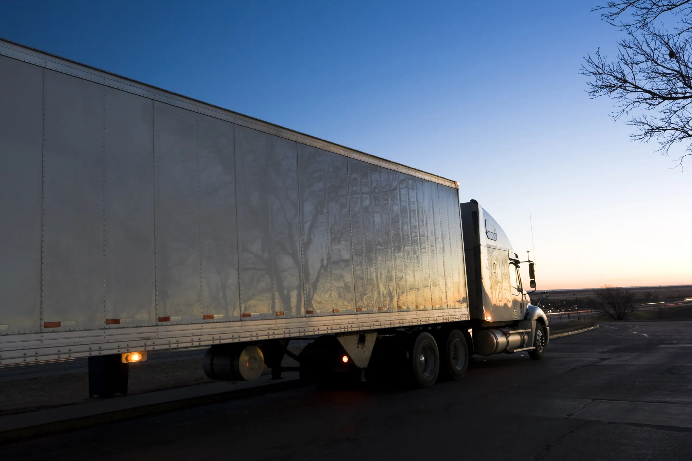 A tractor-trailer at dawn under a deep blue sky, running lights on and a band of sunrise on the horizon.