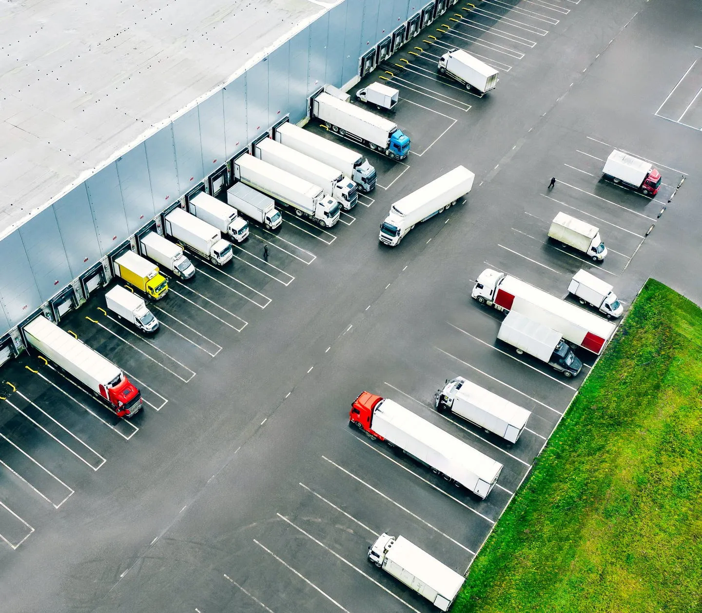 An aerial view of a freight terminal: tractor-trailers backed into a line of loading dock doors, with more rigs and dry van trailers parked across the lot.