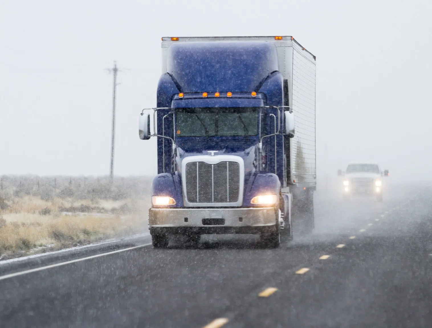 Transport truck hauling a dry van through a snow squall on a rural North American highway