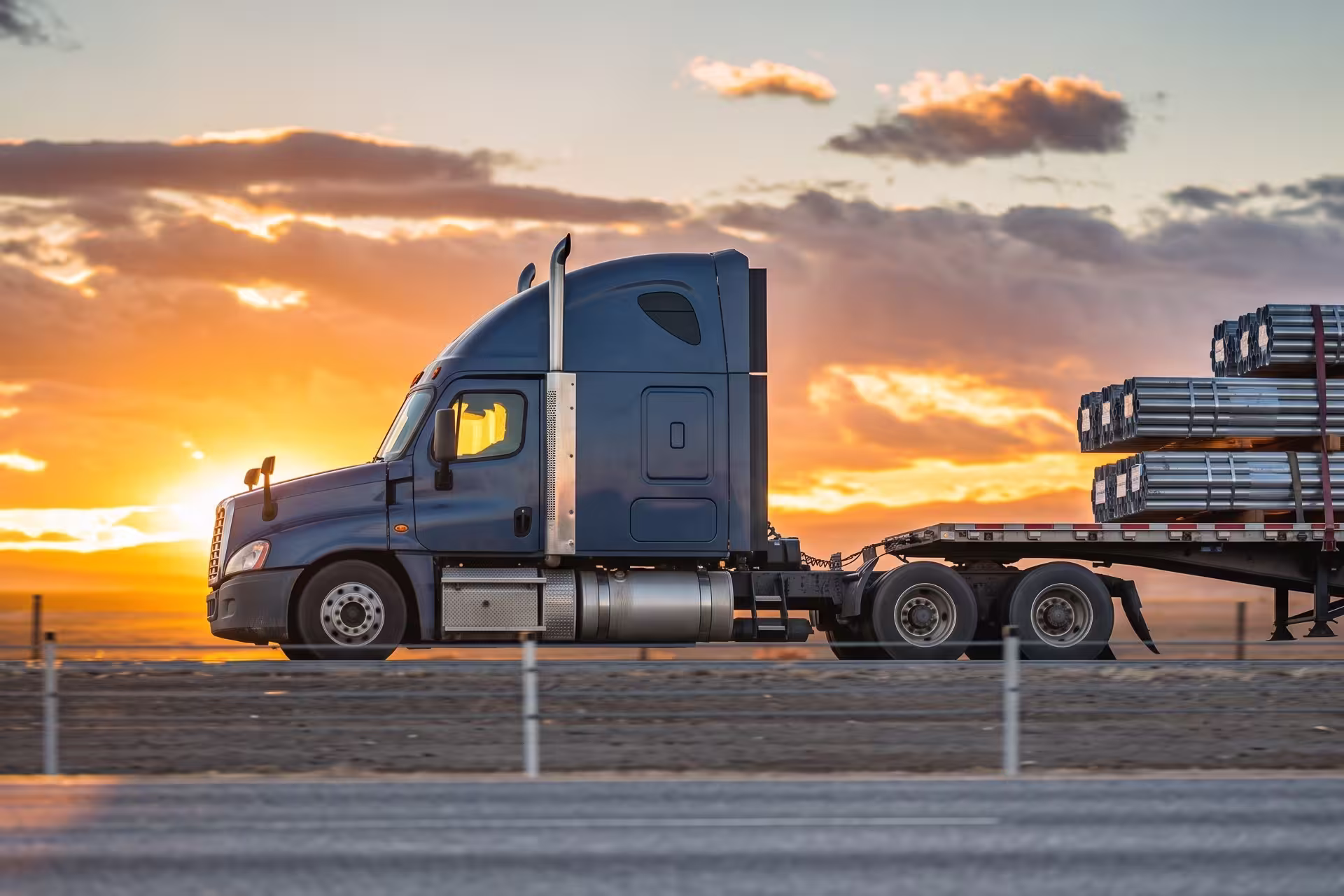 Semi-truck hauling a flatbed trailer loaded with steel pipes at sunset