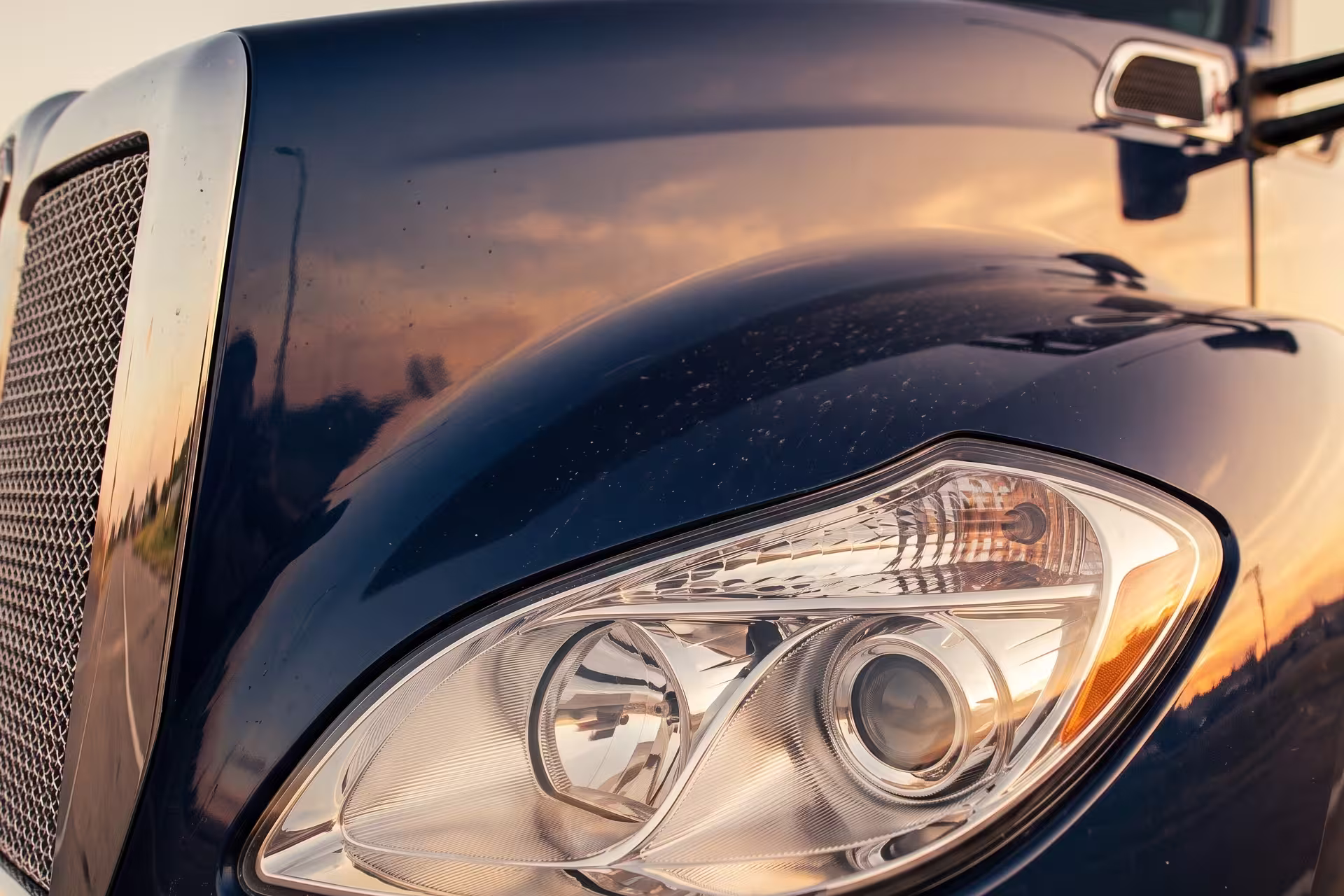 Close-up of a semi-truck's headlight and hood reflecting a sunset sky
