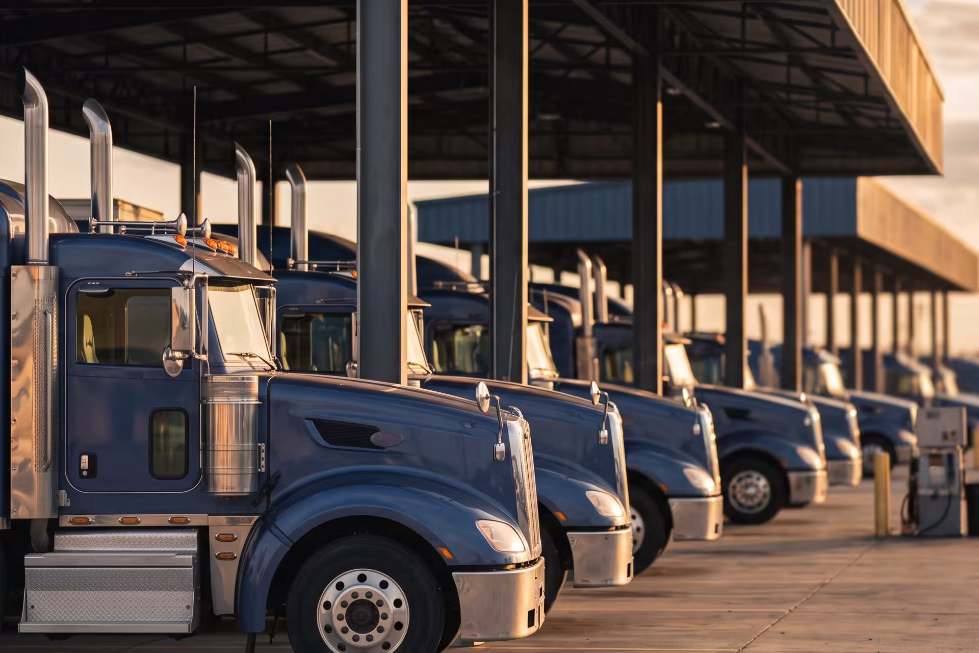 Row of semi-trucks parked side by side under a covered fleet yard canopy at sunset