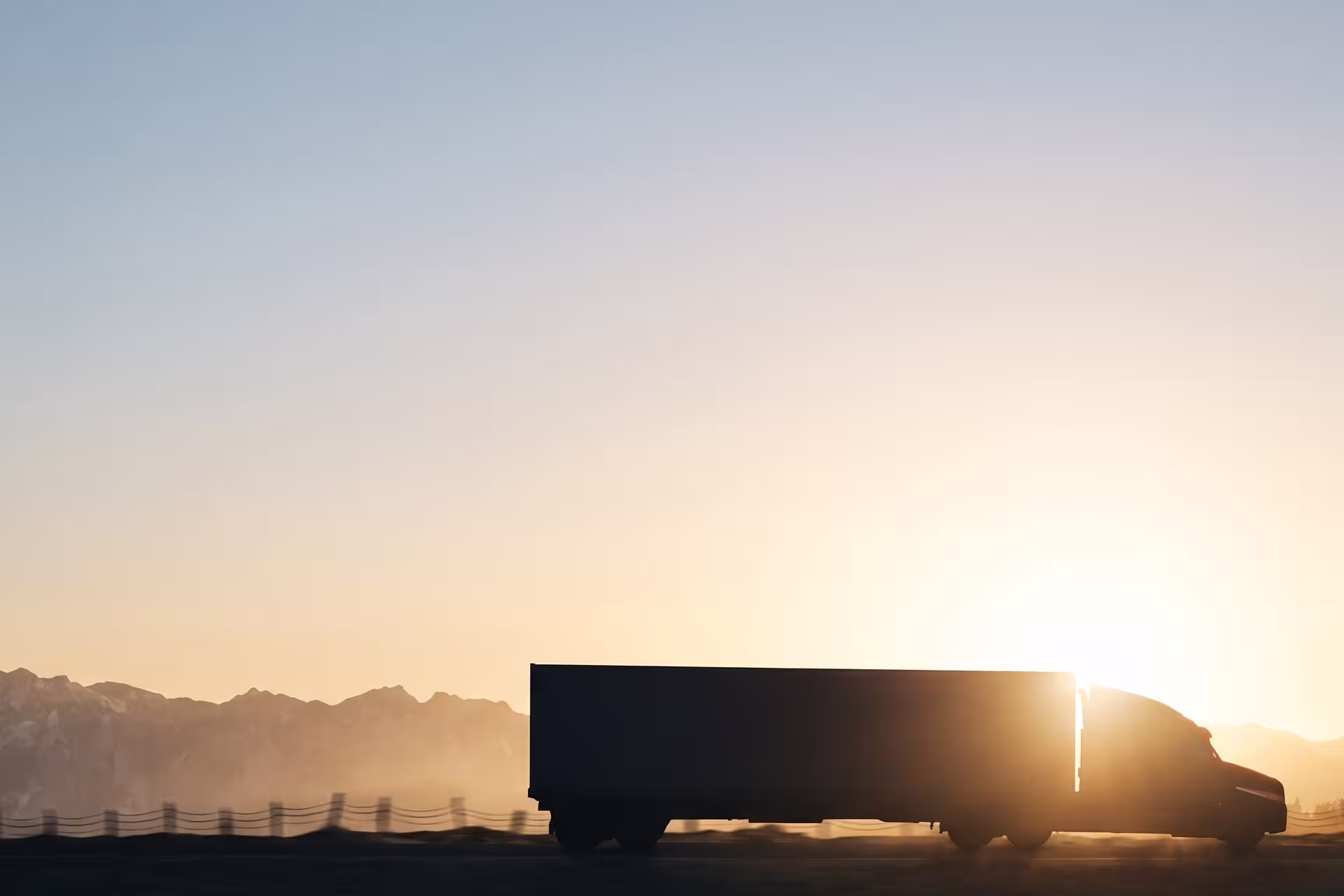 Silhouette of a semi-truck driving past mountains at sunrise