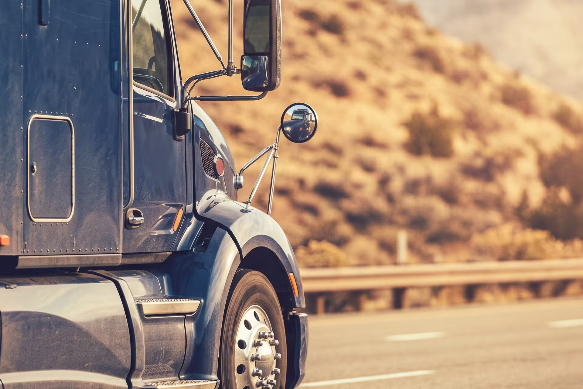 Close-up side view of a semi-truck cab on a desert highway with mountains beyond