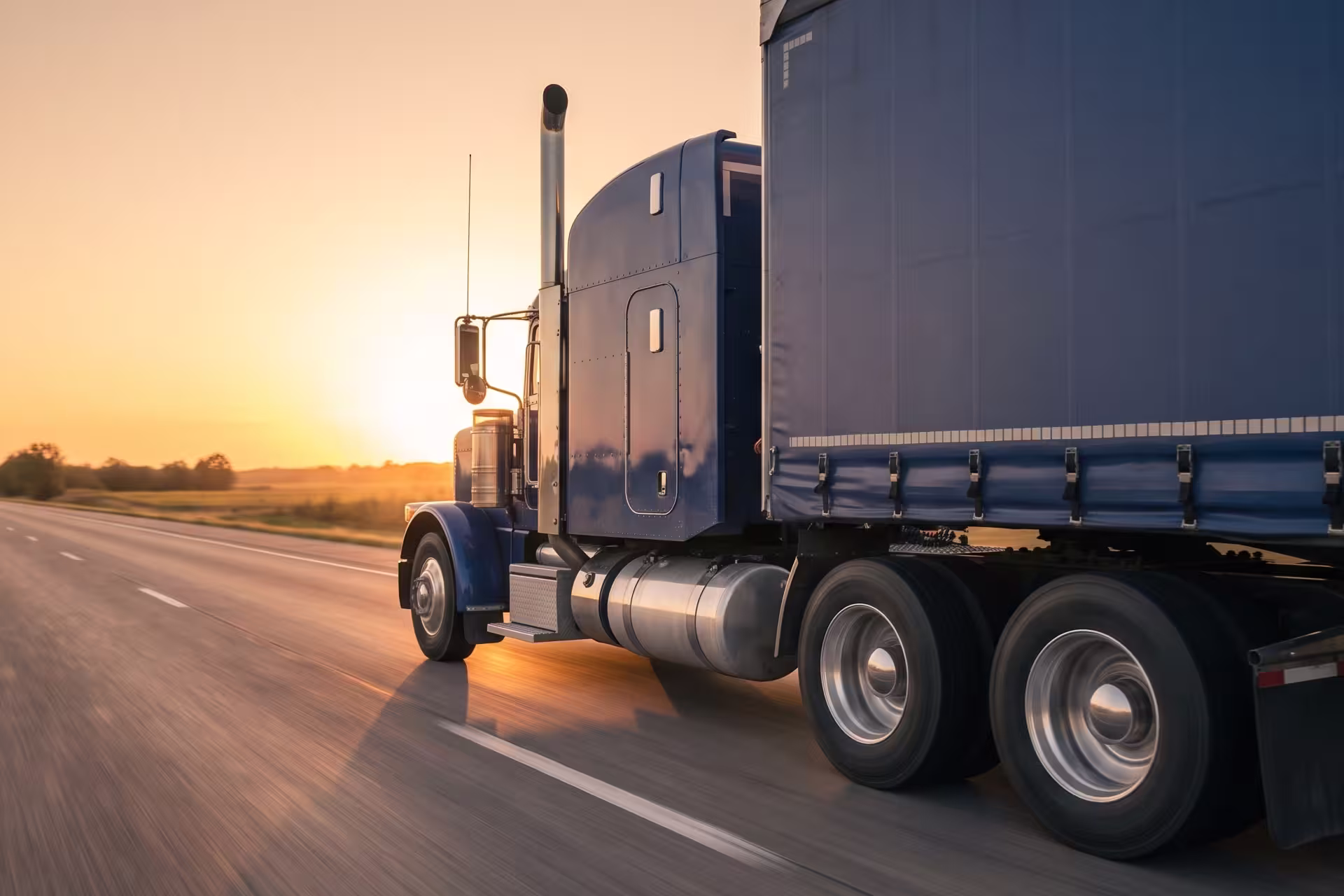 Side view of a semi-truck and trailer driving down a rural highway at sunset