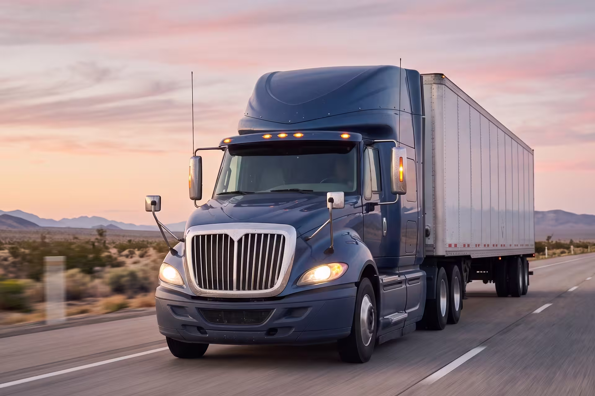 Semi-truck pulling a trailer on a desert highway at dusk with mountains in the background