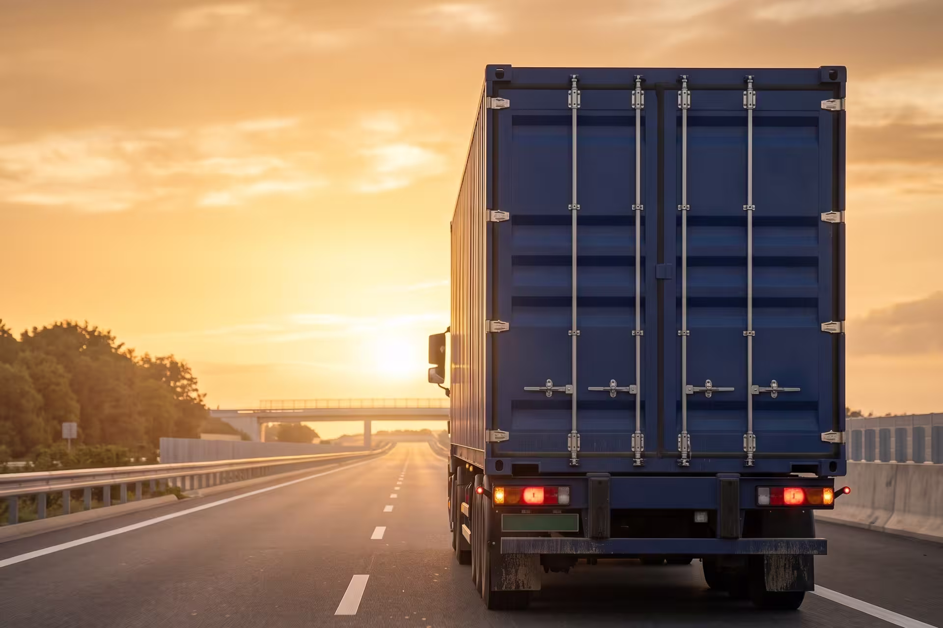 Rear view of a dark blue semi-truck and trailer driving away on a highway at sunset