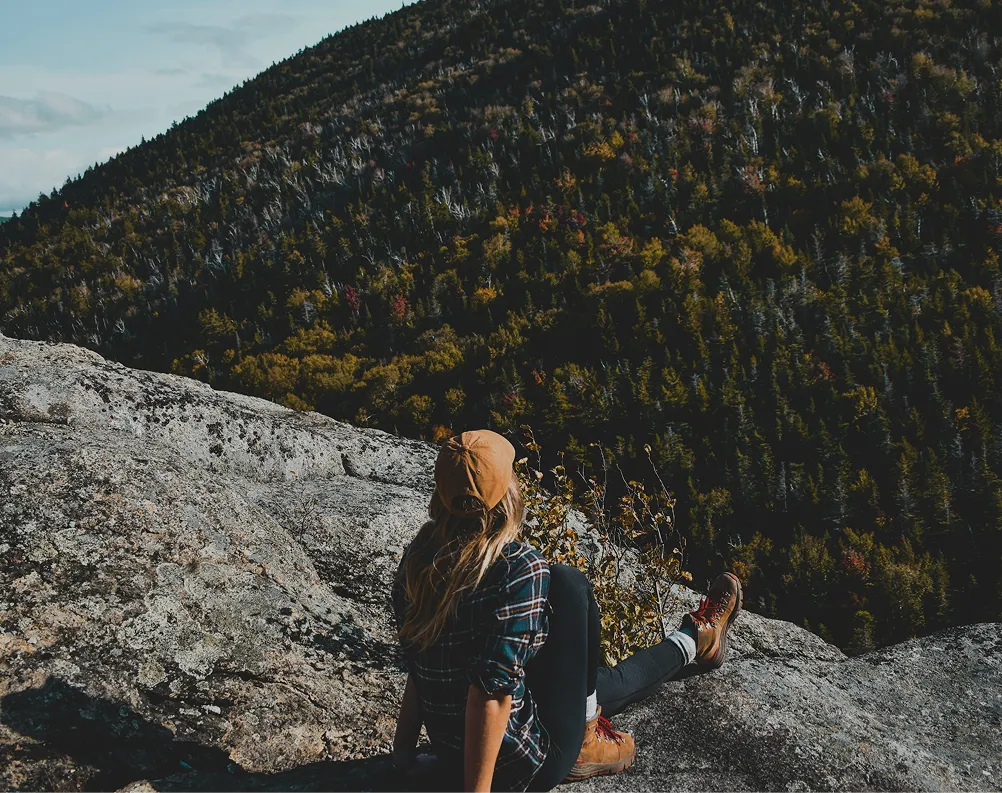 Person in a brown cap and hiking boots sits on a rock, overlooking a forested mountain.