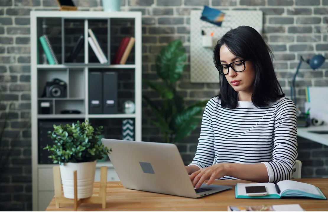 Woman wearing glasses typing on laptop at a wooden desk with notebook and plant in a modern office.