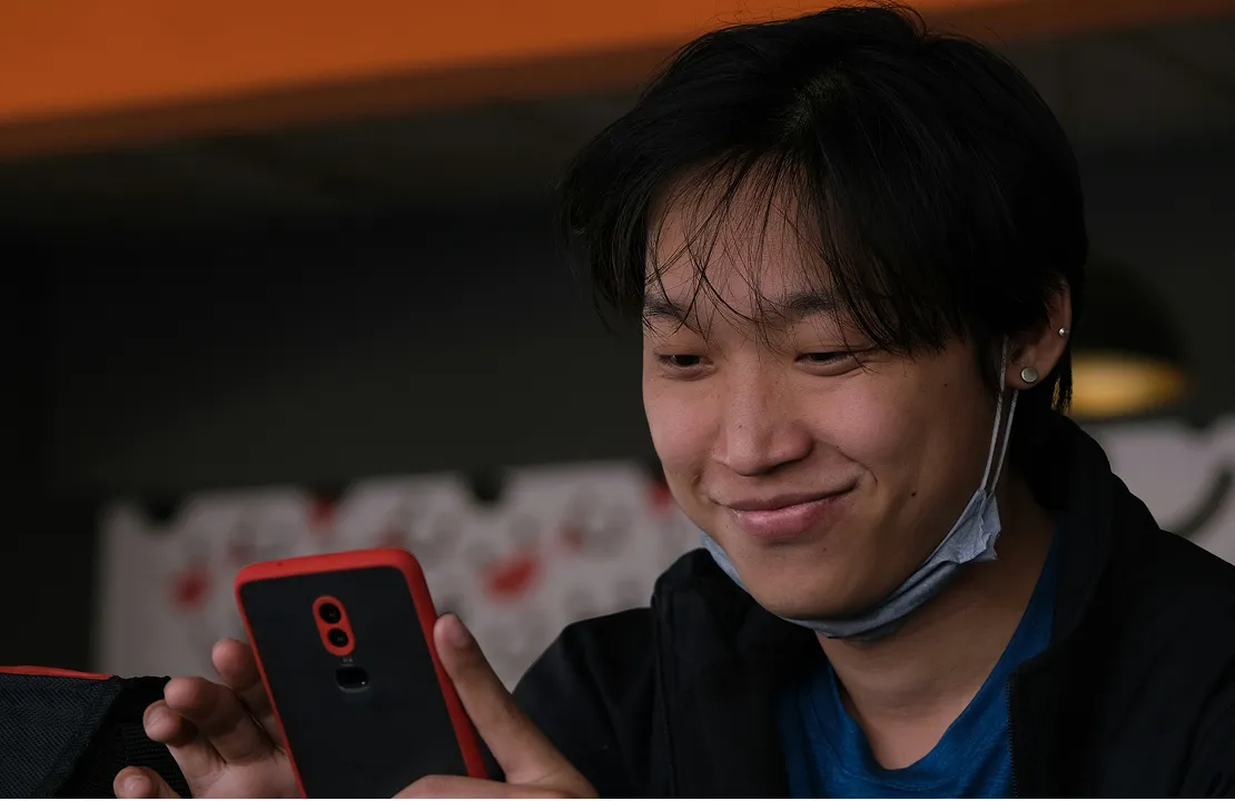 Young person smiling while looking at a smartphone with a red and black case, wearing a mask under the chin.