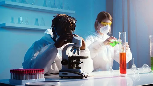 Two scientists in PPE, one using microscope, one pouring green liquid into tall container with red liquid.