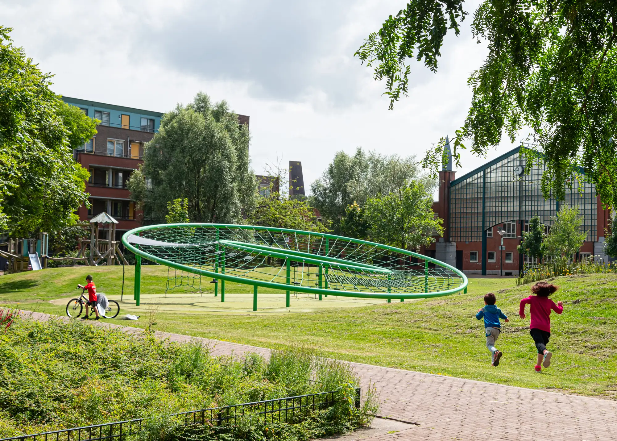 Twee kinderen rennen door een park naar een groot ovaal klimtoestel van groen staal en netten, met een jongen op de fiets en bakstenen appartementen op de achtergrond.