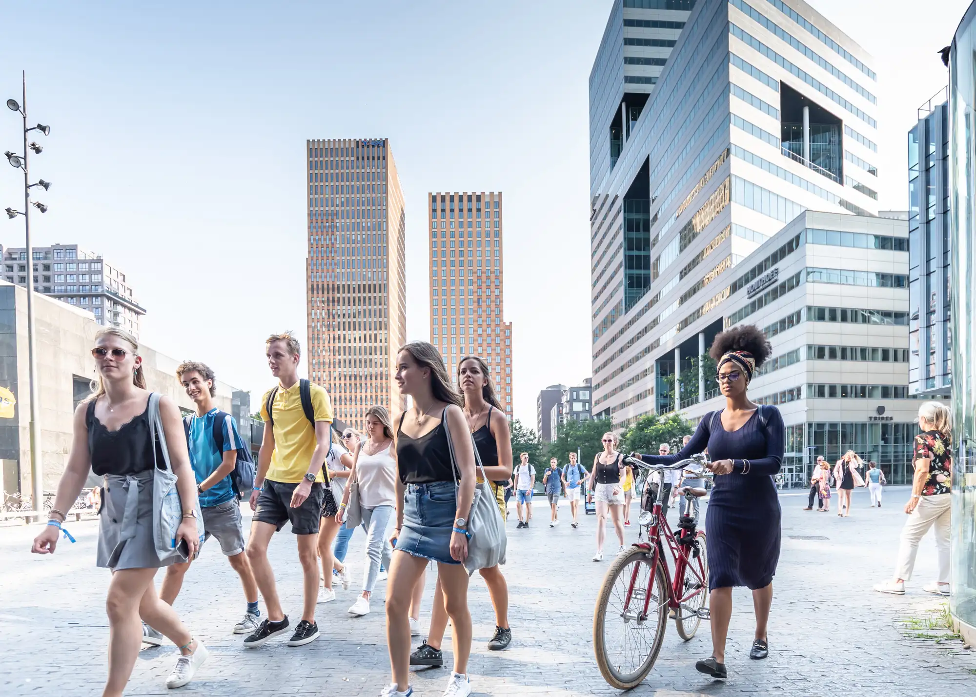 Groep jonge mensen loopt over een plein in de zomer, met een vrouw die haar fiets aan de hand meeneemt, omringd door moderne hoogbouw en kantoortorens.