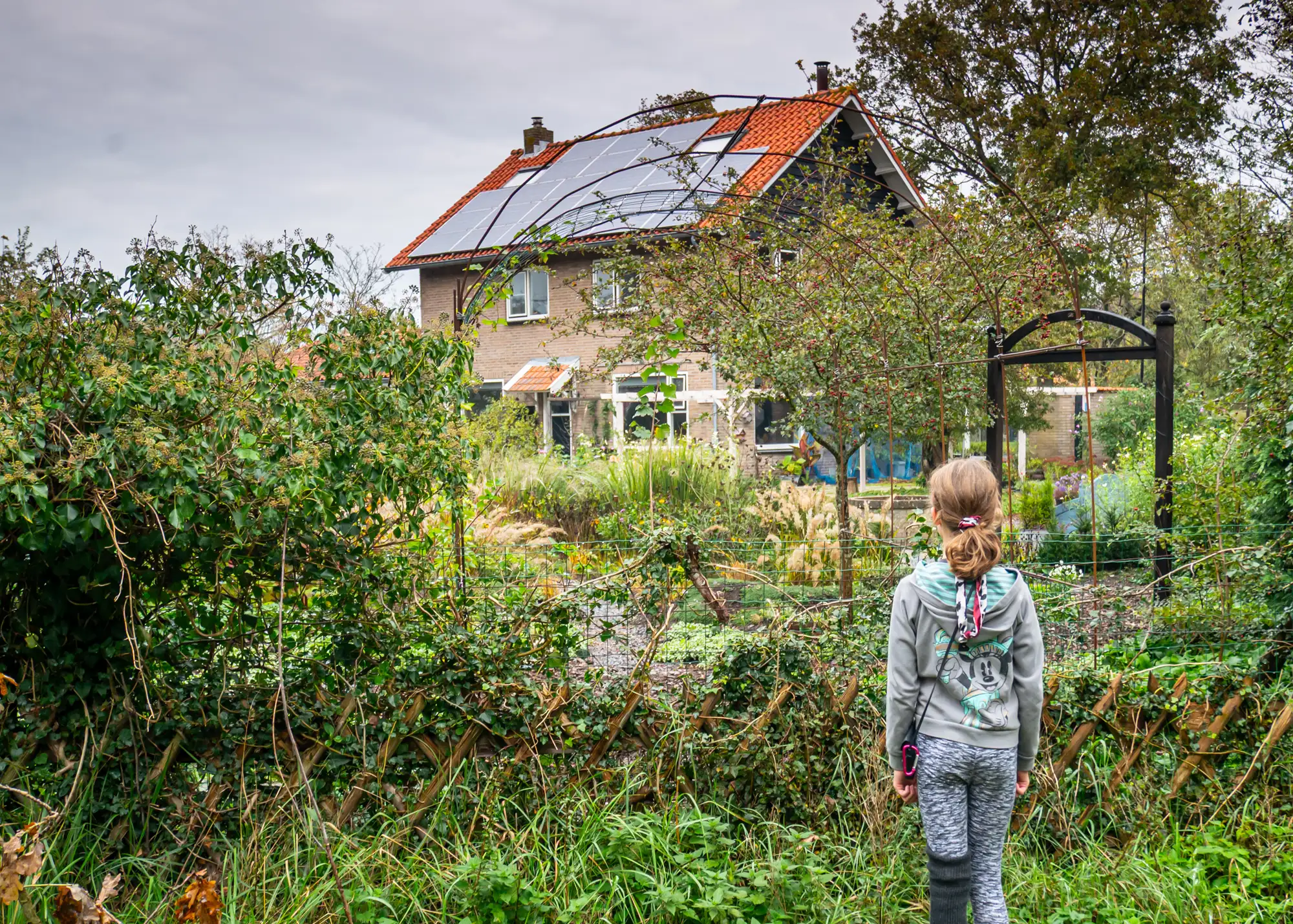 Een meisje van achteren gefotografeerd kijkt over een tuinhek naar een huis met zonnepanelen op het dak, omringd door struiken en planten.