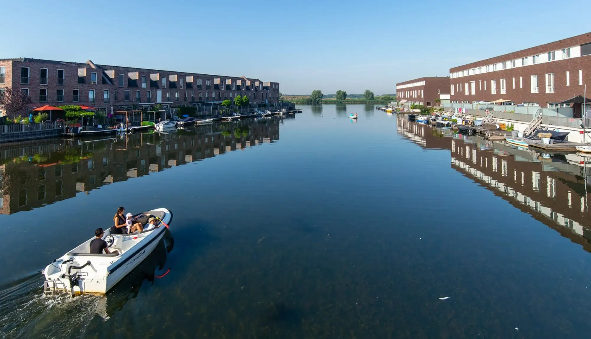 Een motorbootje met gezin vaart over een rustig kanaal tussen twee rijen moderne woningen met aanlegsteigers, het water spiegelt de bebouwing.