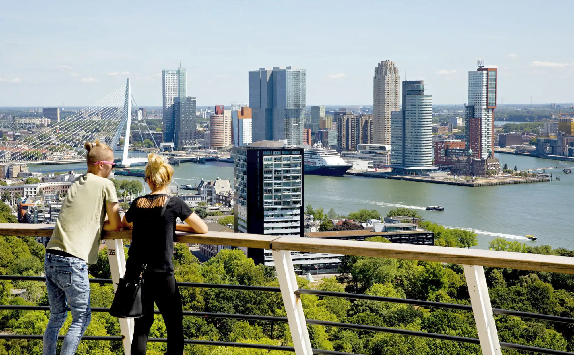 credits: Iris-van-den-Broek. Twee mensen kijken vanaf het uitkijkplatform van de Euromast uit over de Rotterdamse skyline met de Erasmusbrug, de Nieuwe Maas en een cruiseschip aan de Wilhelminakade.