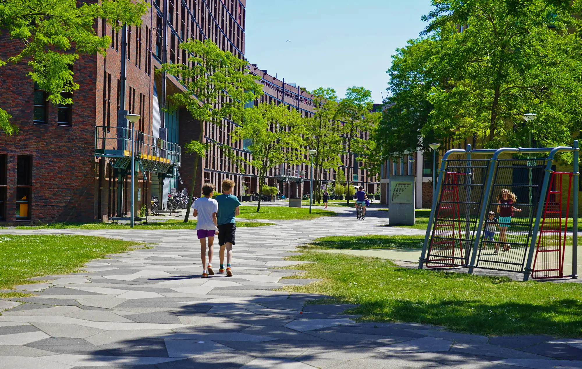 Twee jongens lopen samen over een pad tussen bakstenen appartementengebouwen in een groene woonwijk het funenpark in Amsterdam, met rechts een speeltuin waar kinderen klimmen op een rood-zwart klimrek."