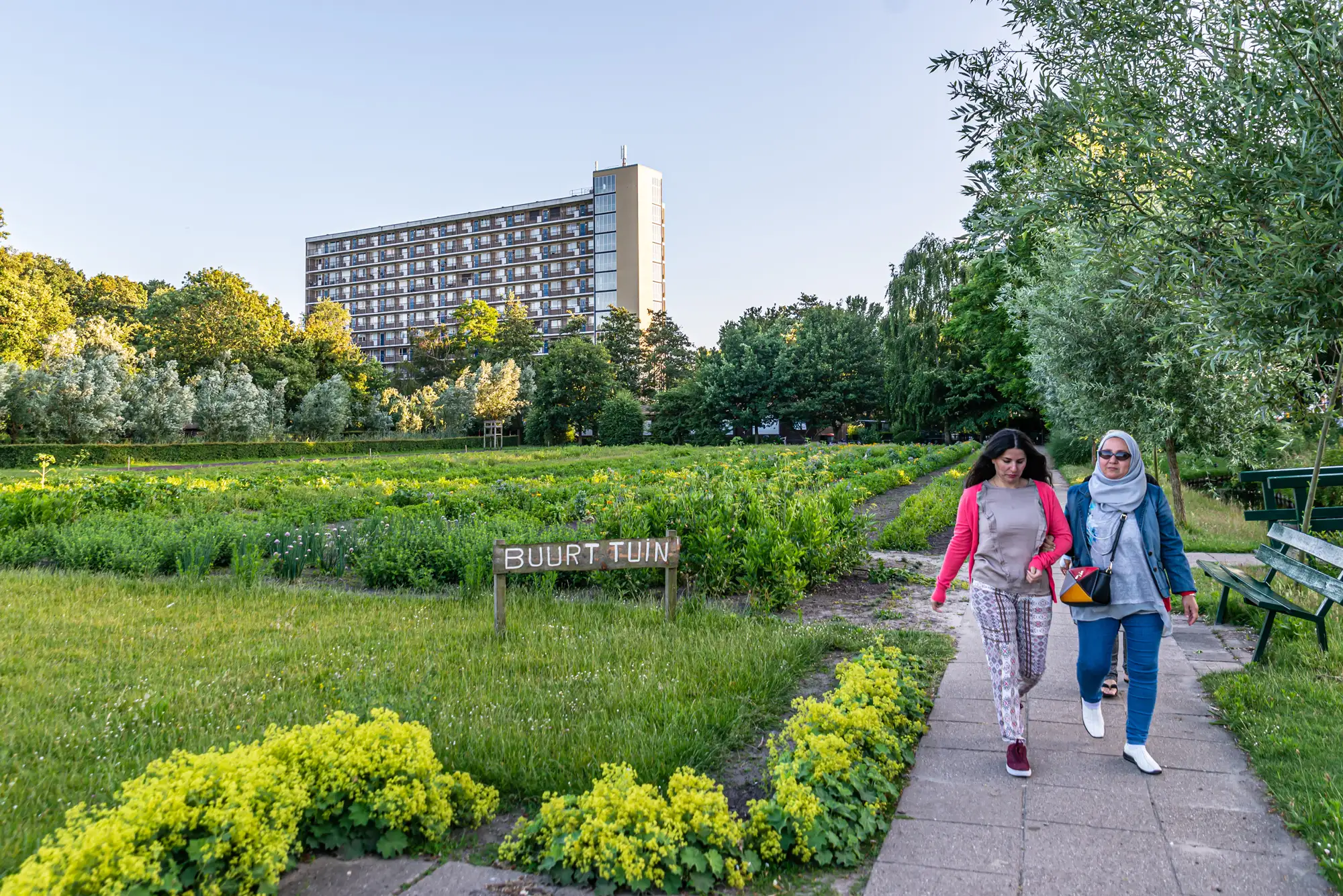Twee vrouwen lopen over een pad langs een weelderige buurtmoestuin met een houten bordje 'Buurt Tuin', met een hoog naoorlogs flatgebouw zichtbaar tussen het groen op de achtergrond.