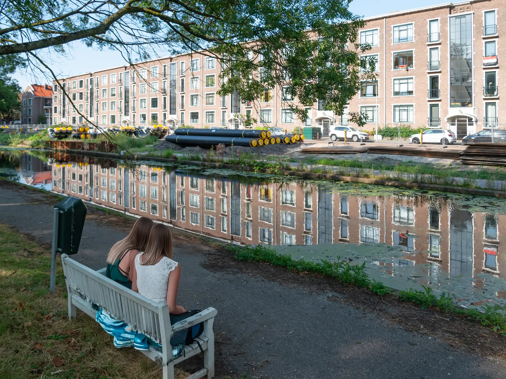Twee meisjes zitten op een bankje langs een kanaal met weerspiegeling van gebouwen in het water.