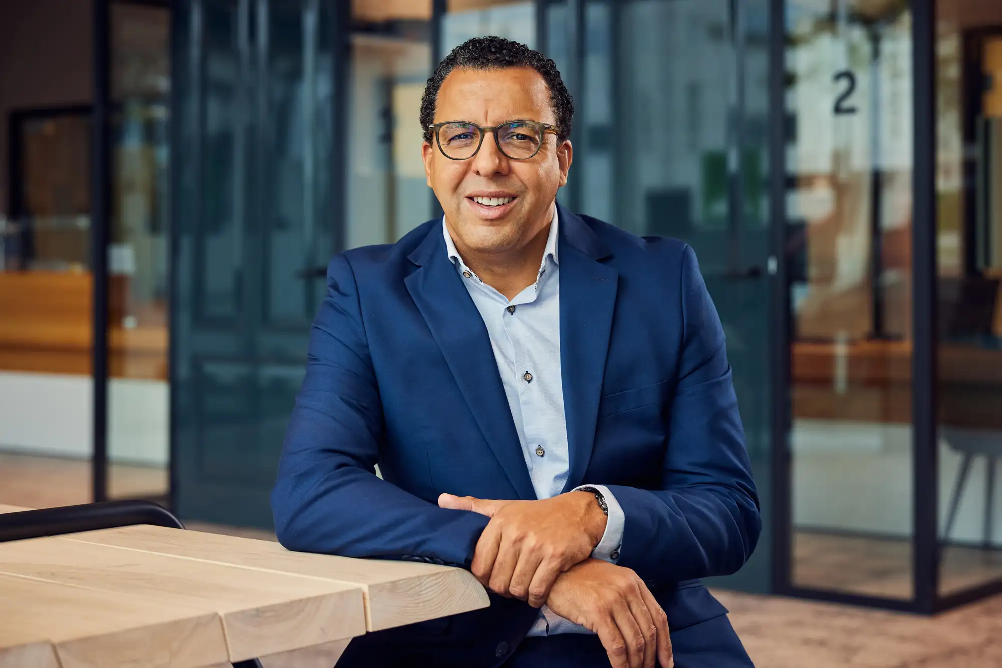 Portretfoto van Mohamed Baba in een donkerblauwe blazer en bril, glimlachend met zijn armen op een houten tafel in een kantooromgeving.
