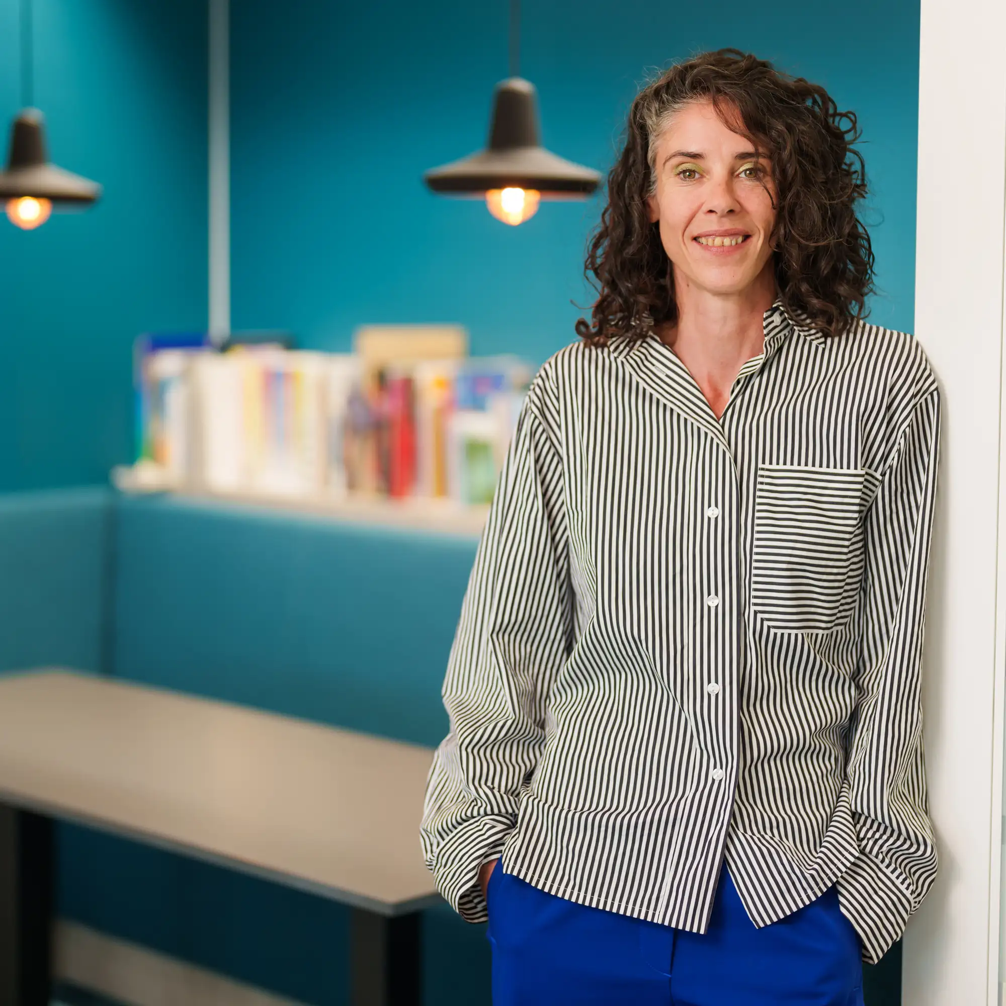 Portretfoto van Anne van Summeren met krullend haar, in een gestreepte blouse, glimlachend in een ruimte met blauwe wand en hangende lampen.
