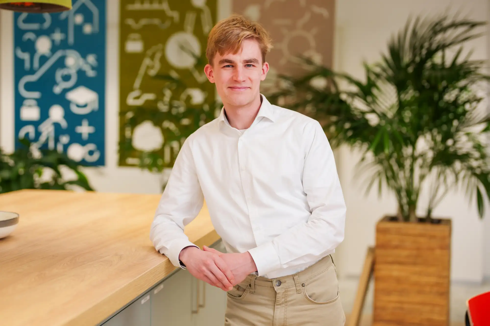 Portretfoto van Joris Dietz in een wit overhemd, leunend tegen een tafel in een kantoorruimte met kleurrijke wanddecoratie en een kamerplant.