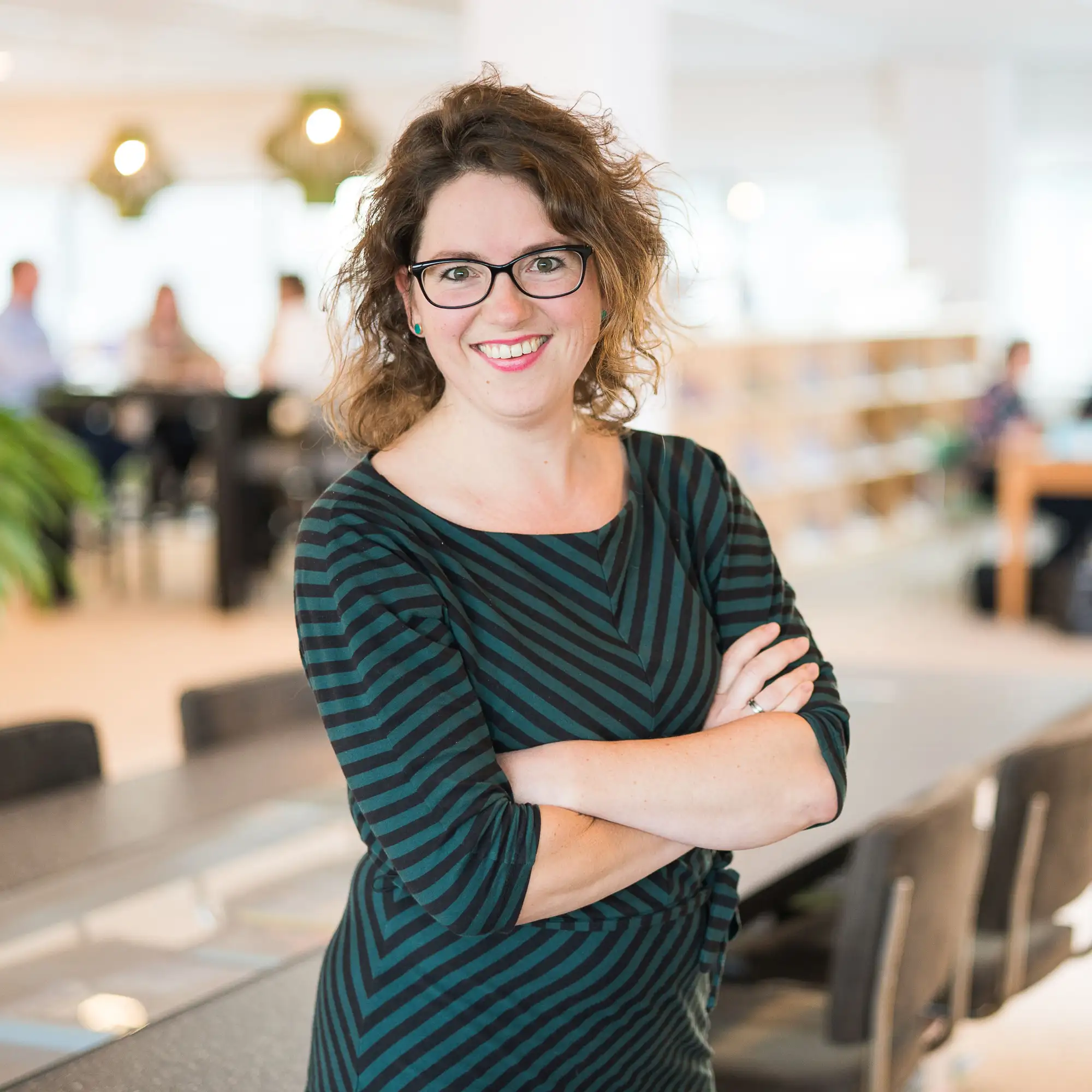 Portretfoto van Janneke met een bril en krullend haar, in een gestreepte jurk en armen over elkaar, glimlachend in een lichte kantoorruimte.