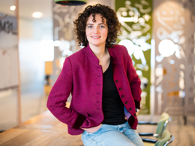 Portretfoto van Susan met krullend bruin haar, in een gemêleerde blazer over een paarse top, glimlachend met een hand in haar zij.