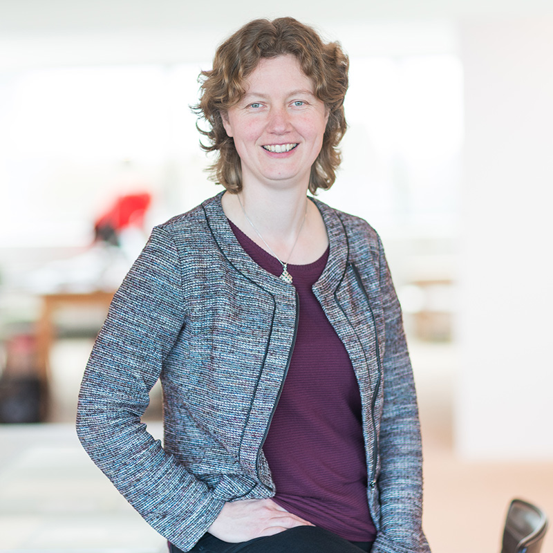 Portretfoto van Susan met krullend bruin haar, in een gemêleerde blazer over een paarse top, glimlachend met een hand in haar zij.