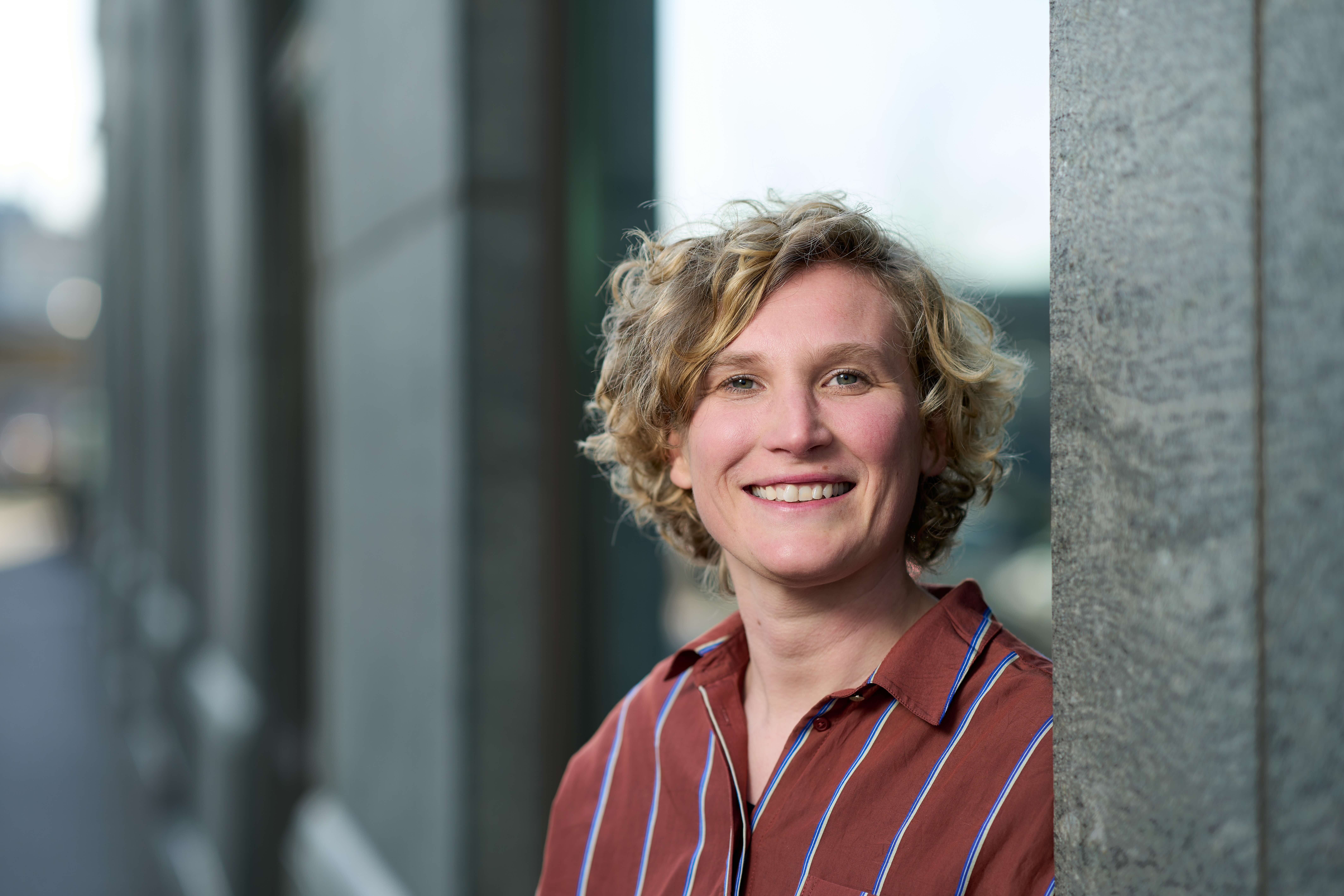 Portretfoto van Susan met krullend bruin haar, in een gemêleerde blazer over een paarse top, glimlachend met een hand in haar zij.
