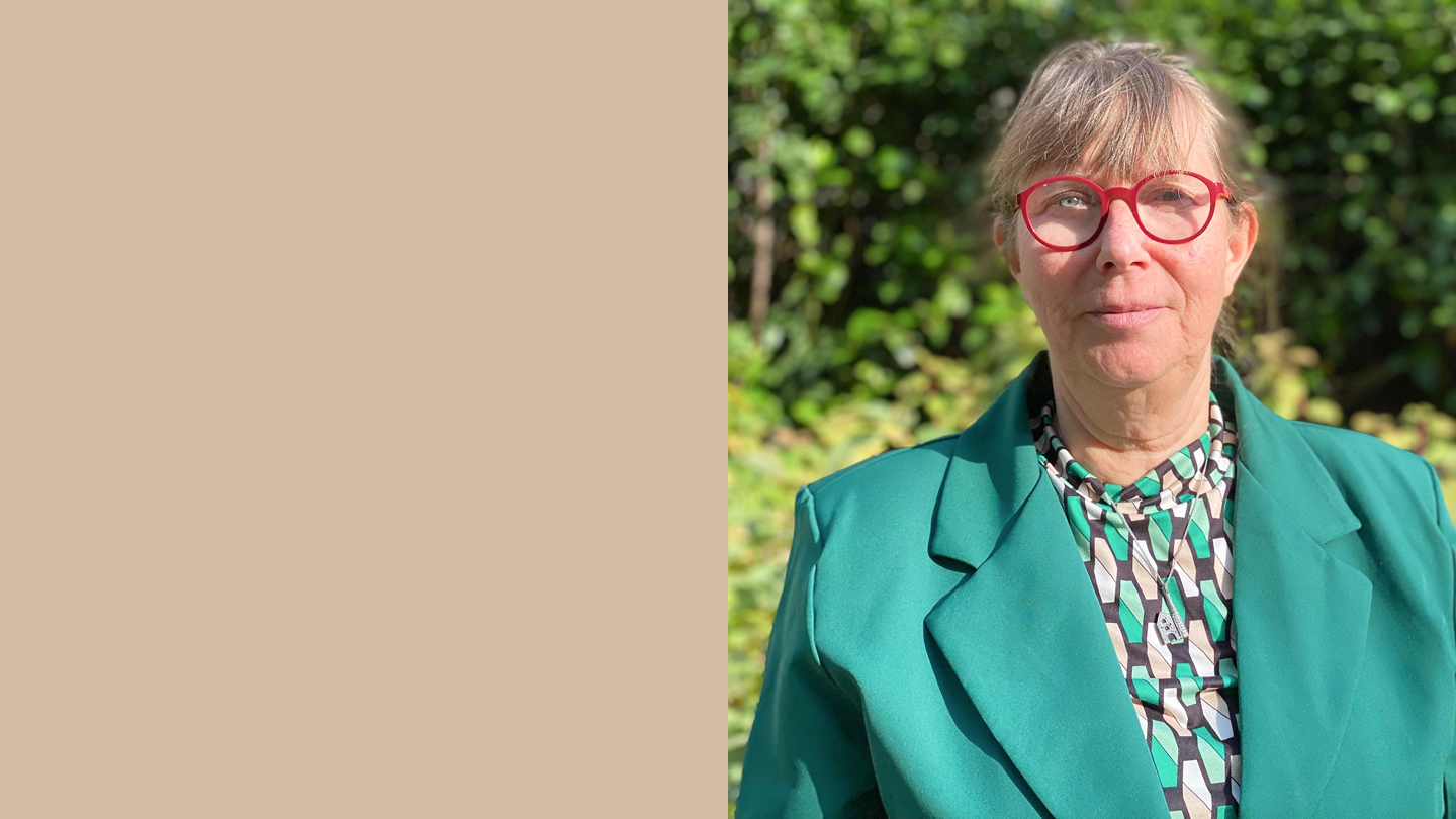 Portretfoto van een vrouw met een bril en groene blazer, voor een achtergrond van groen struikgewas.