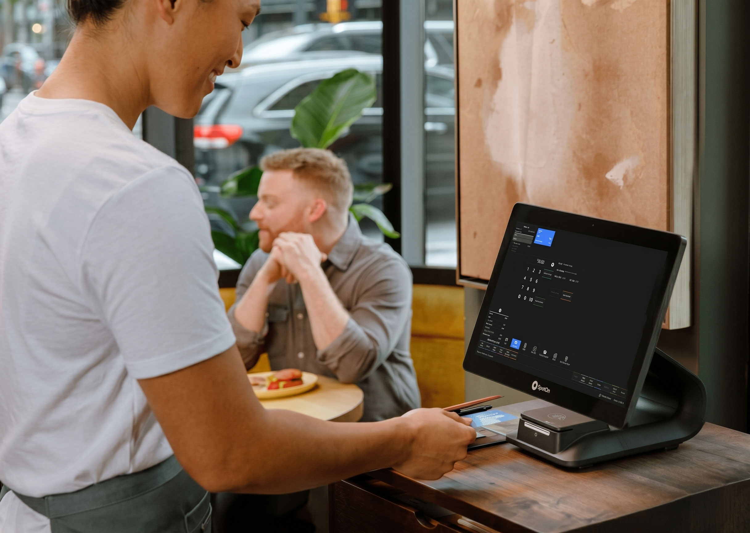 A cafe worker processes a payment on a touchscreen register while a customer waits at a table.