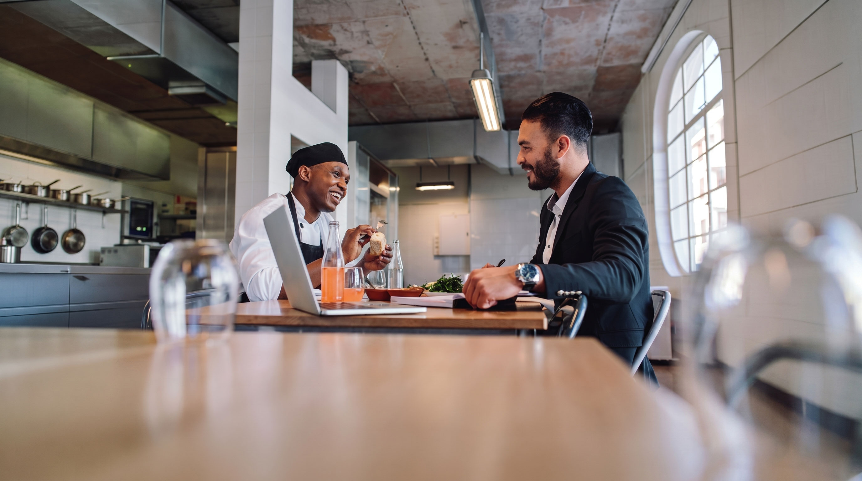 Chef and businessman smiling and discussing over lunch with a laptop and drinks in a kitchen dining area.