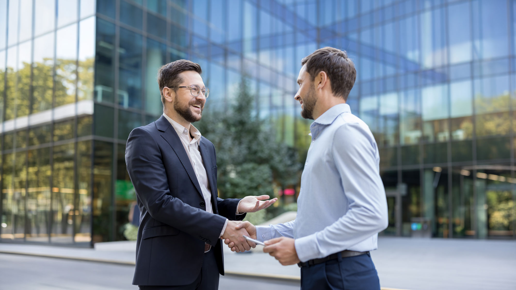Two businessmen shaking hands and smiling outside a modern glass office building.